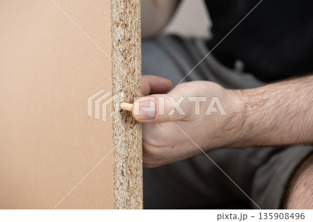 Skilled hand carefully inserting a wooden dowel pin into a pre-drilled hole of a particle board furniture panel, symbolizing DIY assembly and home improvement Skilled hand carefully inserting a wooden dowel pin into a pre-drilled hole of a particle board furniture panel, symbolizing DIY assembly and home improvement 135908496