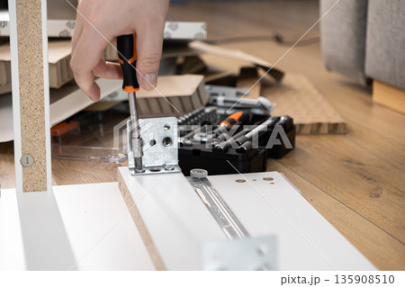 Person's hand skillfully uses a screwdriver to fasten a metal bracket onto white particle board, symbolizing practical furniture assembly and home improvement project 135908510