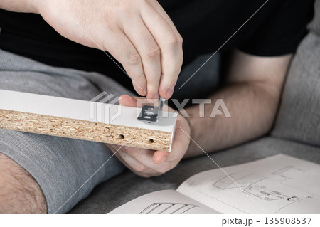 Close-up shows male hands precisely assembling modern white flat-pack furniture, fastening a durable metal bracket with a small hex tool 135908537
