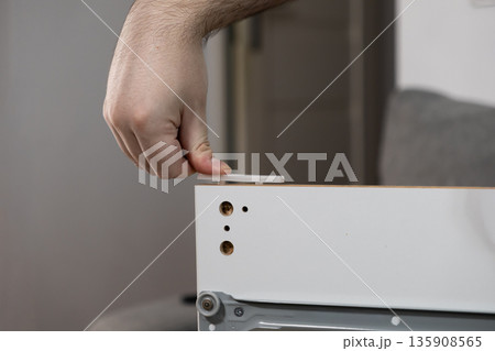Person's hand carefully inserting a white plastic furniture dowel into a pre-drilled hole in a cabinet panel, depicting modern DIY assembly and home improvement 135908565