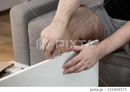 Male hands meticulously assembling modern white flat-pack furniture at home, symbolizing do-it-yourself efforts, new home setup, and personal achievement Male hands meticulously assembling modern white flat-pack furniture at home, symbolizing do-it-yourself efforts, new home setup, and personal achievement 135908573