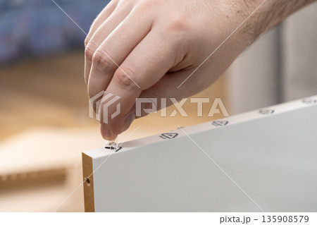 Human hand carefully peeling transparent protective film from white furniture board displaying clear assembly guide symbols, reflecting DIY project effort Human hand carefully peeling transparent protective film from white furniture board displaying clear assembly guide symbols, reflecting DIY project effort 135908579