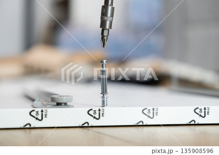 Screwdriver bit aligning to fasten a furniture connector screw into a white flat-pack board, symbolizing DIY assembly and home improvement project Screwdriver bit aligning to fasten a furniture connector screw into a white flat-pack board, symbolizing DIY assembly and home improvement project 135908596
