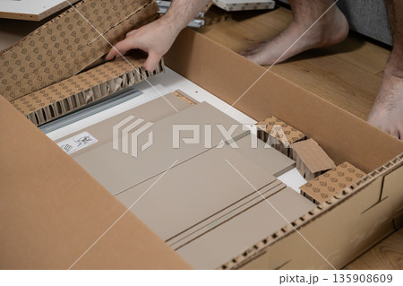 Person's hands unpacking a cardboard box filled with beige flat-pack furniture components, signifying home assembly or a do-it-yourself project Person's hands unpacking a cardboard box filled with beige flat-pack furniture components, signifying home assembly or a do-it-yourself project 135908609