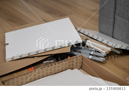 Disassembled flat-pack furniture components, including white panels and hardware, arranged on a wooden floor, signifying home assembly project and DIY effort 135908611
