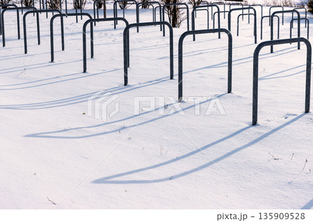 Snow covered bicycle parking area with empty metal bike rack casting long shadows in bright winter sunlight. 135909528