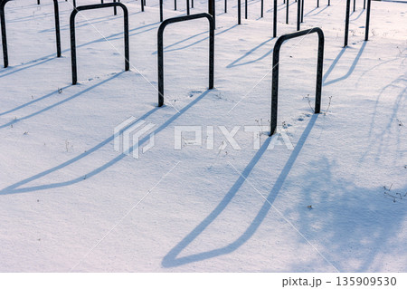 Snow covered bicycle parking area with empty metal bike rack casting long shadows in bright winter sunlight. 135909530
