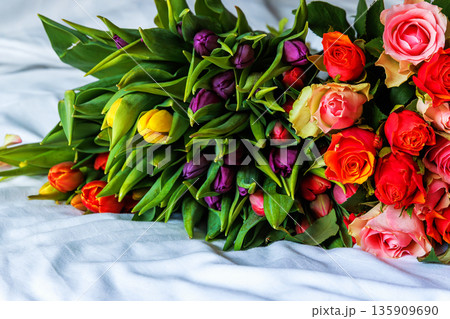 Two vibrant floral bouquets of tulips and roses lying on a bed blanket in a bedroom 135909690