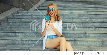 Stylish modern happy smiling young woman with phone in city park, girl holding smartphone on street Stylish modern happy smiling young woman with phone in city park, girl holding smartphone on street 135909734