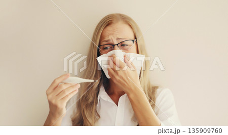 Sick woman sneezing blow nose with tissue, measuring checking body temperature looks at thermometer Sick woman sneezing blow nose with tissue, measuring checking body temperature looks at thermometer 135909760
