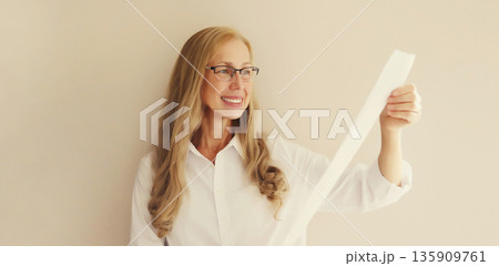 Surprised happy mature woman in glasses looking amazed at the high price tag on long paper receipt 135909761