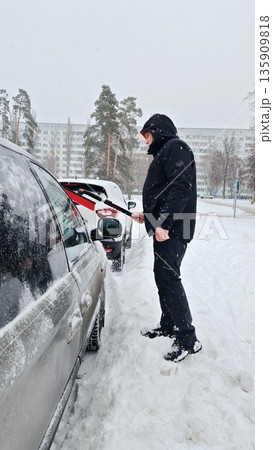 Man Cleaning Snow from Car in Winter. Person using snow brush to clean snow off beige minivan during heavy snowfall in city yard. 135909818