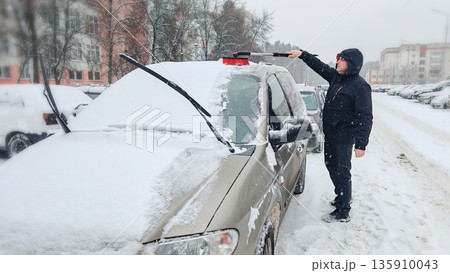 Man Cleaning Snow from Car in Winter. Person using snow brush to clean snow off beige minivan during heavy snowfall in city yard. Man Cleaning Snow from Car in Winter. Person using snow brush to clean snow off beige minivan during heavy snowfall in city yard. 135910043