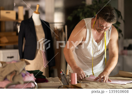 Middle-aged male dressmaker drawing on paper in sewing workshop Middle-aged male dressmaker drawing on paper in sewing workshop 135910608