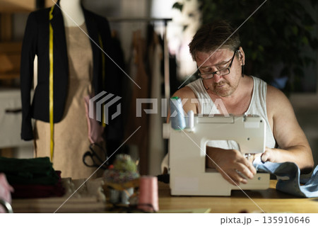 Middle-aged male dressmaker working with sewing-machine at night Middle-aged male dressmaker working with sewing-machine at night 135910646