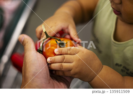 Simple Child Grasps Tomato From Mom, A heartwarming photo of small fingers reaching red fruit in soft kitchen light, for family nutrition or tender moment documentary. 135911509