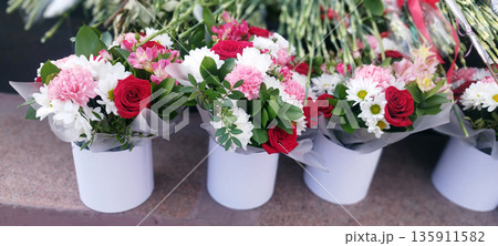 Several vibrant bouquets featuring red roses, pink carnations, white daisies, and alstroemeria are displayed in matching white cylindrical vases outdoors Several vibrant bouquets featuring red roses, pink carnations, white daisies, and alstroemeria are displayed in matching white cylindrical vases outdoors 135911582