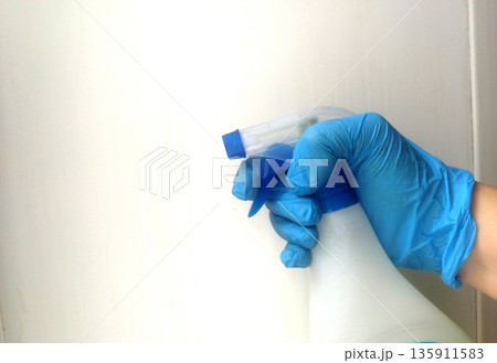 Hand wearing a bright blue disposable glove firmly grips the trigger mechanism of a white spray bottle against a simple, light background, ready for cleaning tasks 135911583