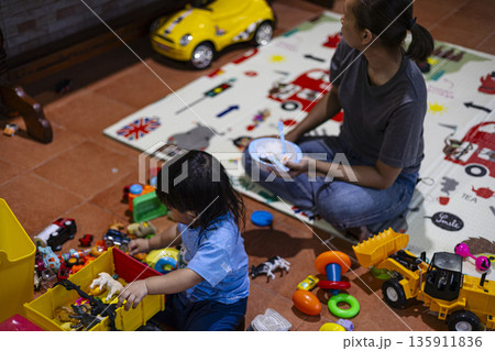 Toddler Playing With Toys While Being Fed By Mother, A premium ad image of mom feeding toddler amid toys, soft light on happy faces, great for nutrition brand or professional care documentary. 135911836
