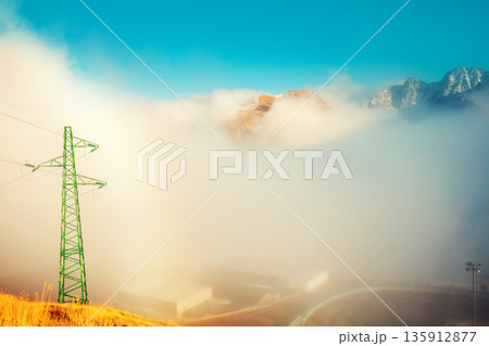 Mountains in clouds. Alpine winding highway above the clouds. Power transmission line in the mountains. Transmission facilities at sunset. Pyrenees, Andorra, Europe Mountains in clouds. Alpine winding highway above the clouds. Power transmission line in the mountains. Transmission facilities at sunset. Pyrenees, Andorra, Europe 135912877