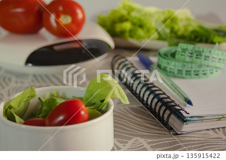 Diet planning essentials with fresh garden vegetables, a kitchen scale, measuring tape, and notebook for tracking health and wellness goals, promoting a balanced lifestyle 135915422