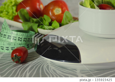 Fresh salad in a bowl on a kitchen scale with a measuring tape and ripe vegetables, illustrating concepts of diet, healthy living, wellness, and weight management 135915425