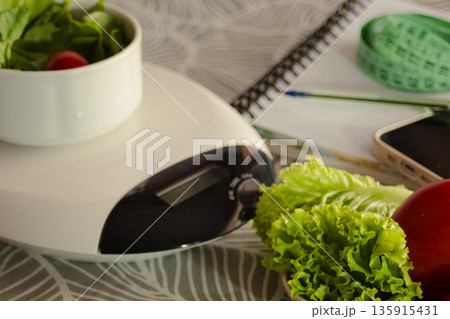 Kitchen objects preparing a healthy diet, with a bowl of fresh salad on a scale, measuring tape, notebook, and a phone for tracking weight loss and wellness journey 135915431