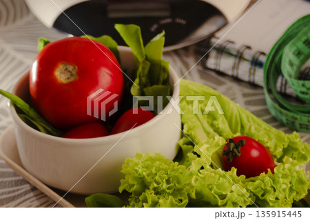 Fresh red tomatoes and crisp lettuce leaves in a ceramic bowl with a kitchen scale, notebook, and measuring tape, symbolizing diet planning, healthy eating habits, and mindful nutrition 135915445