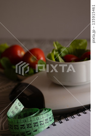 Measuring tape and a notebook resting on a table with a scale in the background. A small bowl of fresh salad and tomatoes creating a concept of healthy living. Diet Measuring tape and a notebook resting on a table with a scale in the background. A small bowl of fresh salad and tomatoes creating a concept of healthy living. Diet 135915461