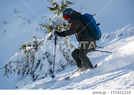 Skier in rugged backcountry snow descending a steep slope with poles and a blue backpack 135922228
