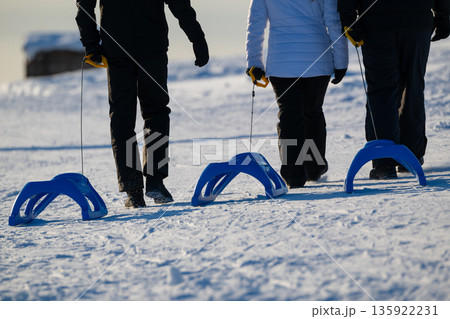 Three people in winter gear walk on a snowy field with blue kick sleds on a bright day 135922231