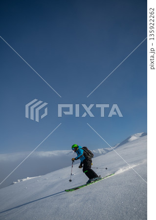 Skier descends a steep snowy slope under a clear blue sky with gear and backpack Skier descends a steep snowy slope under a clear blue sky with gear and backpack 135922262