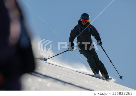 Skier carving down a sunlit snowy slope in protective gear, showcasing focus and speed 135922265