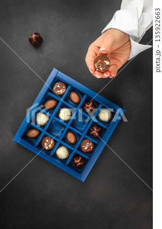 Female hand holding chocolate praline above luxury gift box with assorted chocolates 135922663