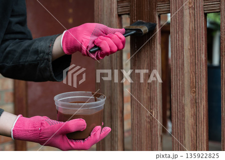 Woman applying wood stain to a wooden fence with a brush on a sunny day in a backyard Woman applying wood stain to a wooden fence with a brush on a sunny day in a backyard 135922825