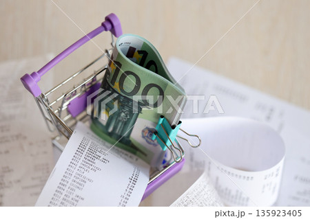 Euro bills bunch in shopping trolley surrounded by many paper receipts close up Euro bills bunch in shopping trolley surrounded by many paper receipts close up 135923405