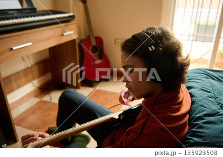 Teenage Boy Wearing Headphones Playing Guitar in Cozy Home Music Room with Keyboard Teenage Boy Wearing Headphones Playing Guitar in Cozy Home Music Room with Keyboard 135923508