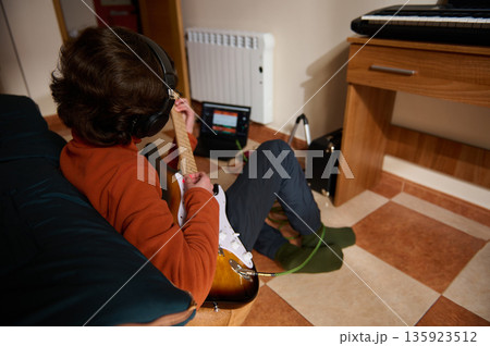 Teenage Boy Playing Electric Guitar With Headphones In Cozy Home Music Setup 135923512