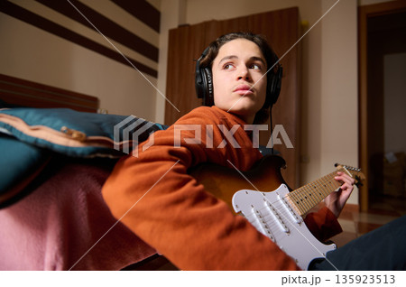 Teenager Playing Electric Guitar With Headphones On Couch In Cozy Room Teenager Playing Electric Guitar With Headphones On Couch In Cozy Room 135923513