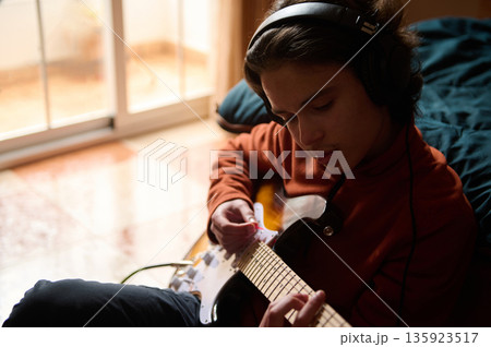 Teenage Boy Playing Electric Guitar With Headphones In Cozy Living Room Teenage Boy Playing Electric Guitar With Headphones In Cozy Living Room 135923517