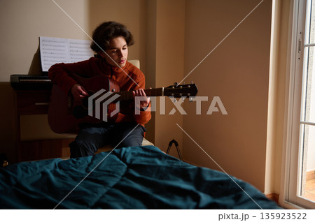 Teen Boy Plays Acoustic Guitar In Bedroom, Focused On Music Hobby 135923522