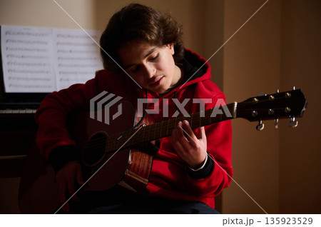 Teenage Boy Playing Acoustic Guitar In A Quiet Room With Sheet Music Backdrop 135923529