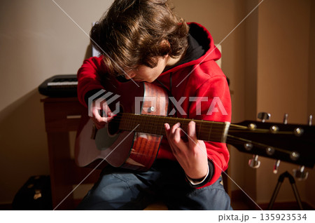 Young Teen Boy In Red Hoodie Playing Acoustic Guitar In A Cozy Home Studio Young Teen Boy In Red Hoodie Playing Acoustic Guitar In A Cozy Home Studio 135923534