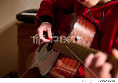 Teen Guitarist Strumming Acoustic Guitar In Red Hoodie At Home Teen Guitarist Strumming Acoustic Guitar In Red Hoodie At Home 135923539