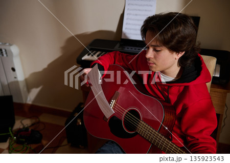Teen Guitar Player Practicing At Home With Sheet Music And Keyboard In Backdrop Teen Guitar Player Practicing At Home With Sheet Music And Keyboard In Backdrop 135923543
