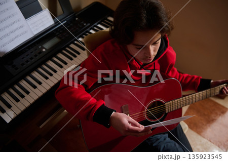 Teenager Plays Guitar While Reading Sheet Music at Home With Keyboard Nearby 135923545