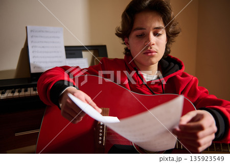 Teen Guitar Player Focused On Sheet Music With Piano In Background Teen Guitar Player Focused On Sheet Music With Piano In Background 135923549