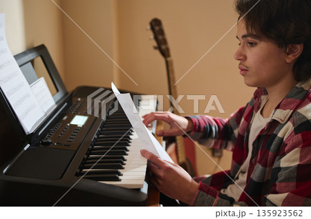 Teenage Boy Plays Keyboard at Home with Sheet Music and Guitar in Background 135923562