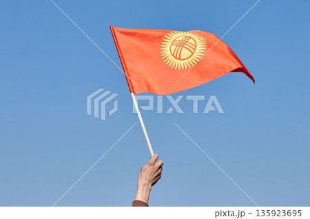 Man waving national flag from Kyrgyzstan against a clear blue sky. Patriotism and national pride concept for country celebration. Man waving national flag from Kyrgyzstan against a clear blue sky. Patriotism and national pride concept for country celebration. 135923695
