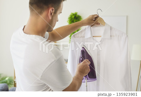 Man ironing white shirt on hanger with steam iron, smoothing wrinkles at home 135923901